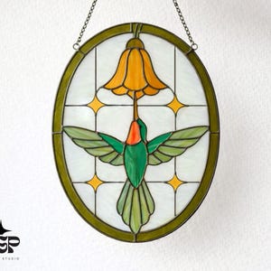 May include: Oval stained glass window art featuring a hummingbird with outstretched wings, a yellow flower, and star accents. The bird has green and orange details, set against a white background with a green border, suspended by a chain.