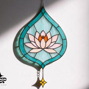 Peut inclure: Vitrail en forme de goutte d'eau avec une fleur de lotus. La fleur est blanche et orange, entourée de verre bleu clair et d'une bordure turquoise. Une petite étoile jaune et une chaîne pendent en bas.
