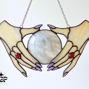 May include: Stained glass artwork depicting two hands cradling a crystal ball. The hands are pale yellow with purple nails and red gemstone rings. The crystal ball is white and swirled. It hangs from a silver chain.