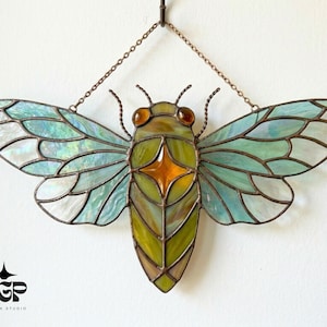 Puede incluir: Adorno de pared de vidriera con forma de cigarra. Alas iridiscentes azules y blancas, cuerpo verde y amarillo, con un diseño de estrella naranja. Se cuelga con una cadena.
