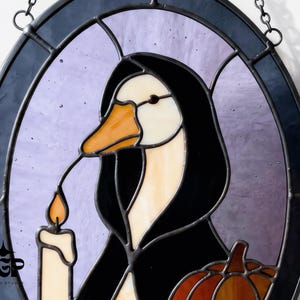 Puede incluir: Vidriera que representa un pato con una capa con capucha negra, sosteniendo una vela con una llama naranja. El pato tiene un pico naranja y est&aacute; sobre un fondo p&uacute;rpura dentro de un marco ovalado negro. Tambi&eacute;n se ve una peque&ntilde;a calabaza.