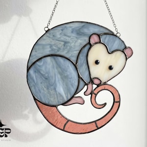 May include: Stained glass opossum suncatcher with a curled tail. The opossum is primarily blue with a cream-colored face and pink ears and tail. The suncatcher is hanging from a silver chain.