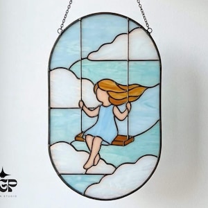May include: Stained glass art piece featuring a girl on a swing amidst clouds. The artwork has a light blue and white color scheme, with a brown swing and the girl's hair in shades of gold. The piece is framed in black and hangs from a chain.