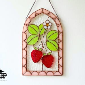 Op de afbeelding: Gebrandschilderd glazen raampaneel met een aardbeienontwerp. Het paneel heeft een roze boogvormig frame en een ophangketting. Het ontwerp bevat twee rode aardbeien, groene bladeren en een witte bloem.