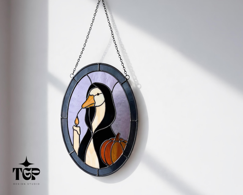 Puede incluir: Vidriera ovalada con un ganso con una capa con capucha negra sosteniendo una vela y una calabaza. La obra de arte est&aacute; enmarcada en un borde azul oscuro y cuelga de una cadena. El fondo es de color p&uacute;rpura claro.