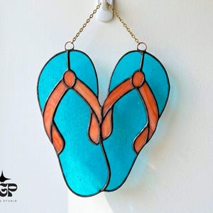 Puede incluir: Obra de arte en vidriera que representa dos chanclas turquesas con correas naranjas, unidas en la parte superior y colgadas de una cadena. El diseño presenta contornos negros sobre un fondo blanco.