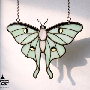 Puede incluir: Un atrapasoles de mariposa de vidrieras en tonos verde pálido, blanco y negro, con un marco de metal color bronce. La mariposa está suspendida por una cadena. El diseño incluye un logotipo que dice "TGP Design Studio".