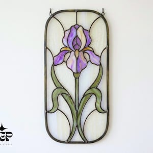 May include: Stained glass panel featuring a purple iris flower with green leaves and a white background. The panel is framed in a dark metal and is designed to hang. The design is in the Art Nouveau style.