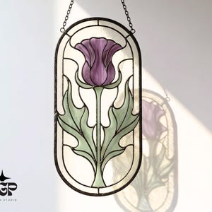 Op de afbeelding: Ovaal glas-in-lood paneel met een paarse distelbloem met groene bladeren en een witte achtergrond. Het paneel is zwart omlijst en hangt aan een ketting.