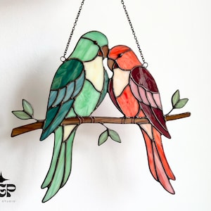 May include: Stained glass art piece featuring two colorful birds perched on a branch. One bird is green and blue, the other is red and pink. The birds are kissing. The piece is suspended by a chain.
