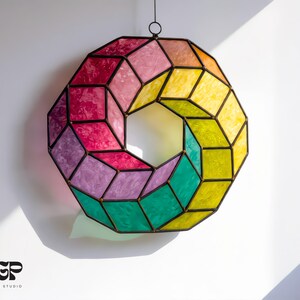 May include: A colorful stained glass geometric wreath with a black metal frame. The wreath features a variety of colors, including pink, purple, yellow, green, and orange. The design is a unique, modern art piece, perfect for home decor.