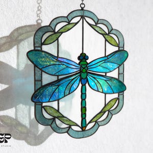 Op de afbeelding: Gebrandschilderde glazen libelle-suncatcher met iriserende blauwe en groene vleugels, een groen lichaam en een decoratief frame. De libelle is gecentreerd in een ovaal frame met groene bladaccenten en een blauwgroene rand. De suncatcher hangt aan een ketting.