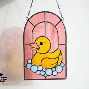 May include: Stained glass window featuring a yellow rubber duck in a bubble bath. The duck has an orange beak and black eyes, set against a pink background. The window is framed in black and hangs from a chain.