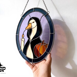 Puede incluir: Vidriera ovalada que representa un ganso con una capa negra, sosteniendo una vela y una calabaza. La obra de arte tiene un borde morado y azul y est&aacute; suspendida por una cadena. La imagen tiene un tema de Halloween.