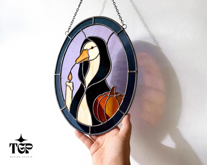Puede incluir: Vidriera ovalada que representa un ganso con una capa negra, sosteniendo una vela y una calabaza. La obra de arte tiene un borde morado y azul y est&aacute; suspendida por una cadena. La imagen tiene un tema de Halloween.