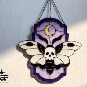 Könnte beinhalten: Glasmalerei-Motten mit Totenkopf-Design, an einer Kette hängend. Die Motte hat weiße und schwarze Flügel, einen Totenkopf und einen schwarzen Körper. Der Hintergrund ist lila mit einem gelben Halbmond.
