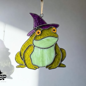 Può includere: Vetrata a forma di rana con cappello da strega viola. La rana è principalmente verde e gialla con occhi arancioni e pancia verde chiaro. La rana è appesa a una corda.