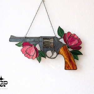 May include: Stained glass art piece depicting a revolver with a wooden handle, adorned with pink and red flowers and green leaves. The gun is suspended by a chain. The piece is a unique decorative item.