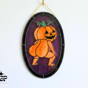 May include: An oval stained glass suncatcher featuring an orange pumpkin character with a jack-o'-lantern face, depicted in a twerking pose against a deep purple background. The piece is framed in black metal and hangs from a gold-colored chain.