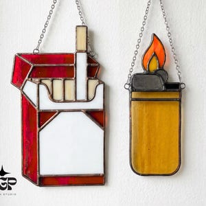 Può includere: Due opere d'arte in vetro colorato appese a catene. Una è un pacchetto di sigarette rosso e bianco, l'altra un accendino giallo con una fiamma arancione e gialla. I pezzi sono realizzati con vetro al piombo.