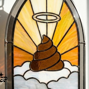 May include: An arched stained glass panel featuring a brown swirled shape centred above white clouds. A yellow and orange sunburst pattern radiates from behind the shape, which is topped with a black wire halo. The TGP Design Studio logo is visible.