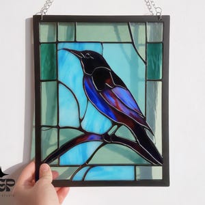 May include: Stained glass panel featuring a bird perched on a branch. The bird has black, blue, and purple feathers. The background is a mix of blue and green glass, framed in black with a silver chain for hanging.
