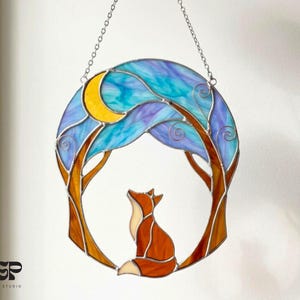 Puede incluir: Vidriera que representa un zorro bajo un árbol y una luna creciente. El diseño circular utiliza colores azul, morado, naranja y amarillo. La pieza está suspendida por una cadena.