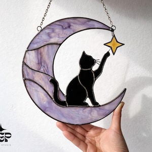 以下が含まれることがあります： 黒猫が黄色の星に手を伸ばすシルエットが特徴の、ステンドグラスの三日月。 月は紫と白の色合いが混ざり合い、チェーンで吊り下げられています。 家の装飾用の装飾品です。