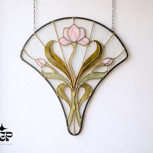 Puede incluir: Un panel de vidrieras en forma de abanico, con una flor rosa y hojas verdes. La obra de arte tiene un marco de metal negro y está suspendida por una cadena. El diseño incluye una flor central, dos capullos y hojas estilizadas.