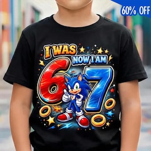 May include: Black t-shirt featuring a colorful graphic of Sonic the Hedgehog with the text "I WAS 6 NOW I AM 7". The design includes the numbers 6 and 7, stars, and rings. The shirt is a birthday-themed item.