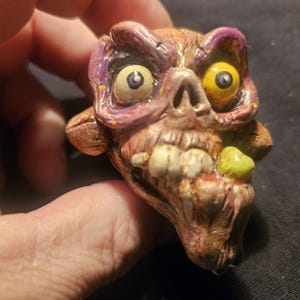 May include: A grotesque, brown, and purple sculpted head with yellow eyes and a gaping mouth. The head has a decaying appearance, with exposed teeth and a greenish growth on the side. The sculpture is held in a hand.