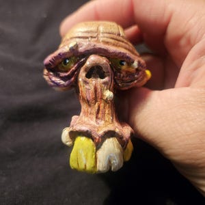 May include: A hand-painted, whimsical sculpture of a creature's head. The sculpture features a brown, textured head with purple accents, green eyes, and a detailed, open mouth. The base has yellow and off-white elements.