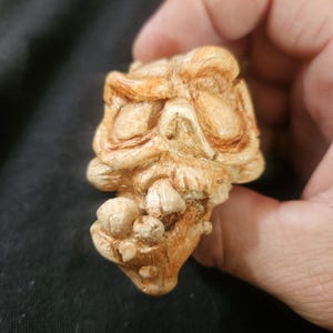 May include: A detailed, tan-colored sculpture of a grotesque face with exaggerated features. The sculpture has a rough texture and appears to be made of a hard material. The face has large, furrowed brows, a prominent nose, and a toothy grin. The overall design is reminiscent of a gargoyle.
