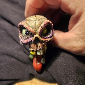 May include: A detailed, hand-painted skull figurine with a grotesque, cartoonish design. The skull features a red tongue, yellow teeth, and purple-rimmed eye sockets. The skull is held against a dark background.