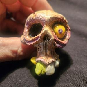 May include: A detailed, hand-painted skull figurine with a grotesque, zombie-like appearance. The skull features a yellow eye with a purple iris, and the teeth are a mix of white and yellow. The color palette includes shades of brown, purple, and green.