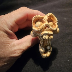 May include: A detailed, tan-colored skull figurine with exaggerated features. The skull has large eye sockets, a prominent jaw, and a textured surface. The figurine is likely made of a hard material and is designed to be a decorative item.