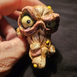 May include: A grotesque, hand-painted figurine of a monster's head with large yellow eyes, a wrinkled face, and yellow accents. The figure is primarily brown and tan with pink and purple highlights, and is held in a hand.