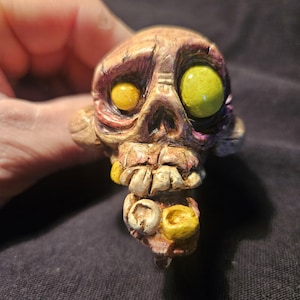 May include: A detailed, hand-painted skull sculpture with yellow and green eyes, and yellow teeth. The skull has a weathered, aged appearance with shades of brown, pink, and purple. The sculpture is a unique piece of art.