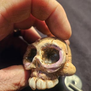 May include: A small, hand-painted skull figurine with a weathered appearance. The skull features a beige base color with purple accents around the eye sockets and white teeth. A metal screw and washer are visible.