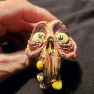 May include: A grotesque, hand-painted figurine of a creature's head. The sculpture features a decaying, textured appearance with yellow eyes, a yellow appendage, and a red spot. The color palette includes shades of purple, brown, and yellow.
