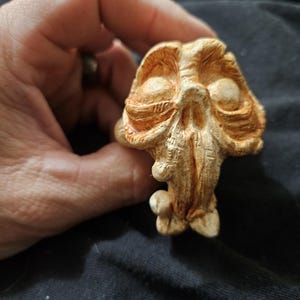 May include: A detailed, tan-colored sculpture of a stylized skull with prominent eye sockets and textured surfaces. The sculpture has a rough, organic appearance, with a small, rounded base.