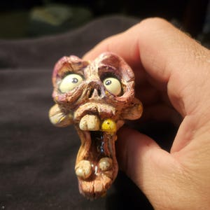 May include: A grotesque, hand-painted zombie head figurine with bulging white eyes, yellow teeth, and a gaping mouth. The sculpture features a detailed, textured surface with shades of brown, purple, and yellow. The figurine is held in a hand against a dark background.