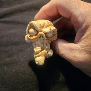 May include: A small, tan-colored sculpture of a stylized skull with exaggerated features. The skull has large eye sockets, a prominent brow, and a detailed, textured surface. The sculpture is held in a hand against a dark background.