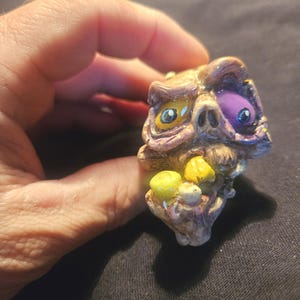 May include: A grotesque, hand-painted ring featuring a zombie-like face with yellow and purple eyes, and yellow and white teeth. The ring is held by a hand against a dark background. The ring is a unique piece of jewelry.