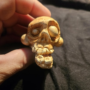 May include: A detailed, beige-colored skull sculpture with prominent eye sockets, a defined nose, and a detailed jawline. The sculpture has a textured surface and is held in a hand, showcasing the intricate craftsmanship of the piece.