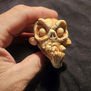 May include: A detailed, hand-painted skull figurine with an aged, weathered appearance. The skull has prominent eye sockets, a detailed nose, and a wide, toothy grin. The color palette includes shades of beige, brown, and orange, giving it a vintage look.