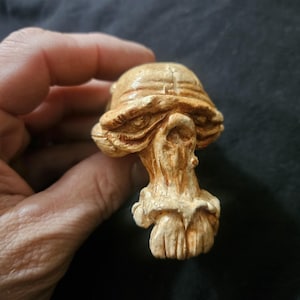 May include: A detailed, hand-carved mushroom sculpture in shades of tan and cream. The mushroom features a textured cap and stem, with intricate details. The sculpture is likely made of wood or a similar material.