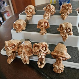 May include: A collection of ten light brown, hand-sculpted skull-shaped drawer knobs with detailed facial features, including prominent eye sockets, noses, and teeth, arranged on a white three-tiered display stand with gray textured surfaces.