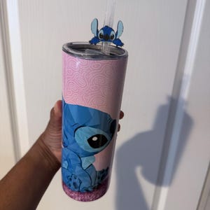 May include: A pink and blue tumbler with a cartoon character design. The tumbler has a pink background with a swirl pattern and a blue character illustration. A blue character straw topper and a clear straw are included.
