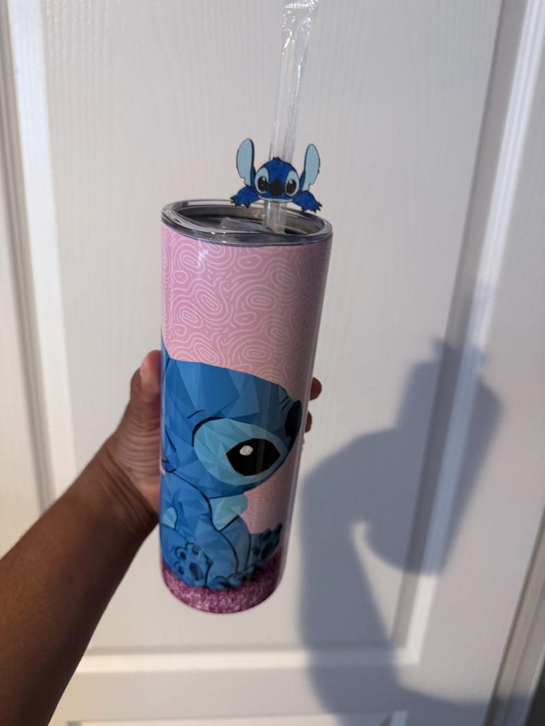 May include: A pink and blue tumbler with a cartoon character design. The tumbler has a pink background with a swirl pattern and a blue character illustration. A blue character straw topper and a clear straw are included.